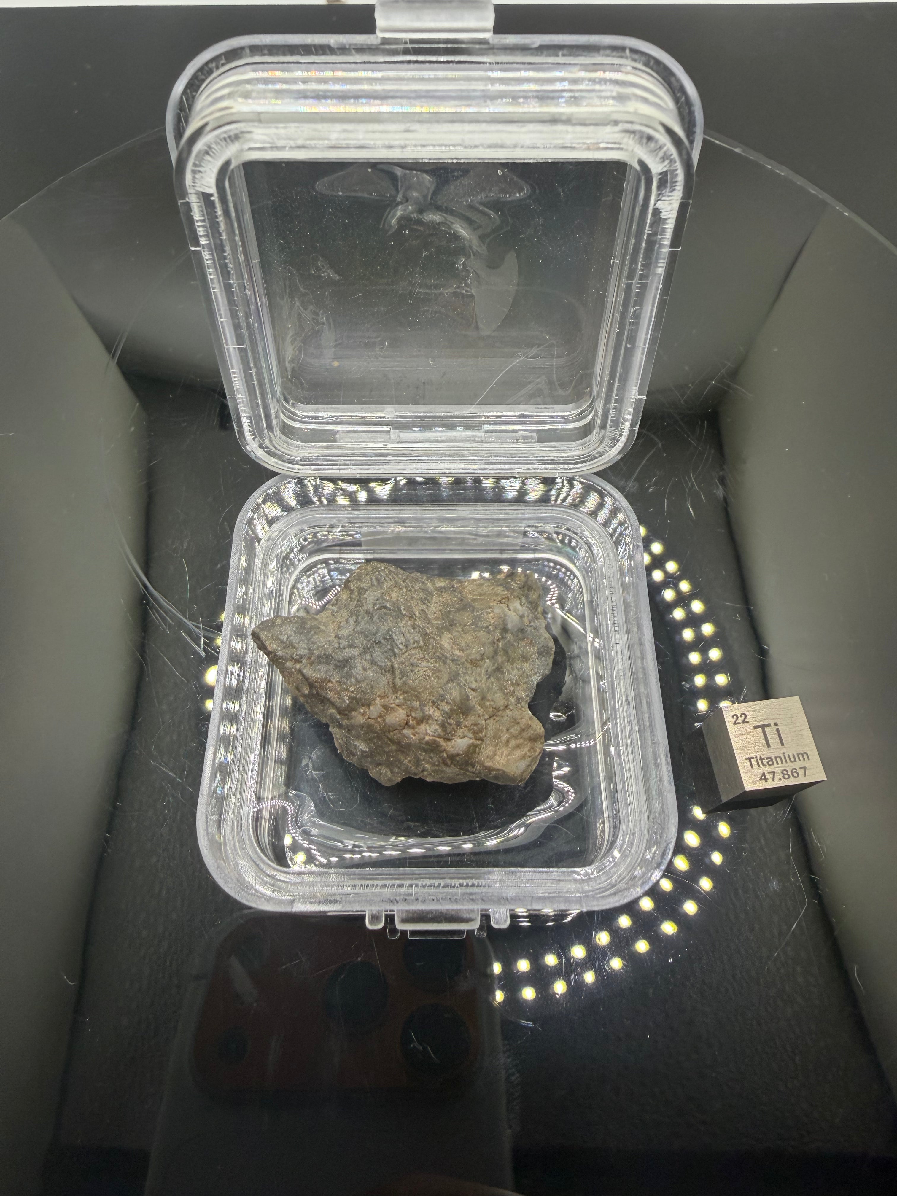 back side of lunar sample in clear plastic case next to scale cube