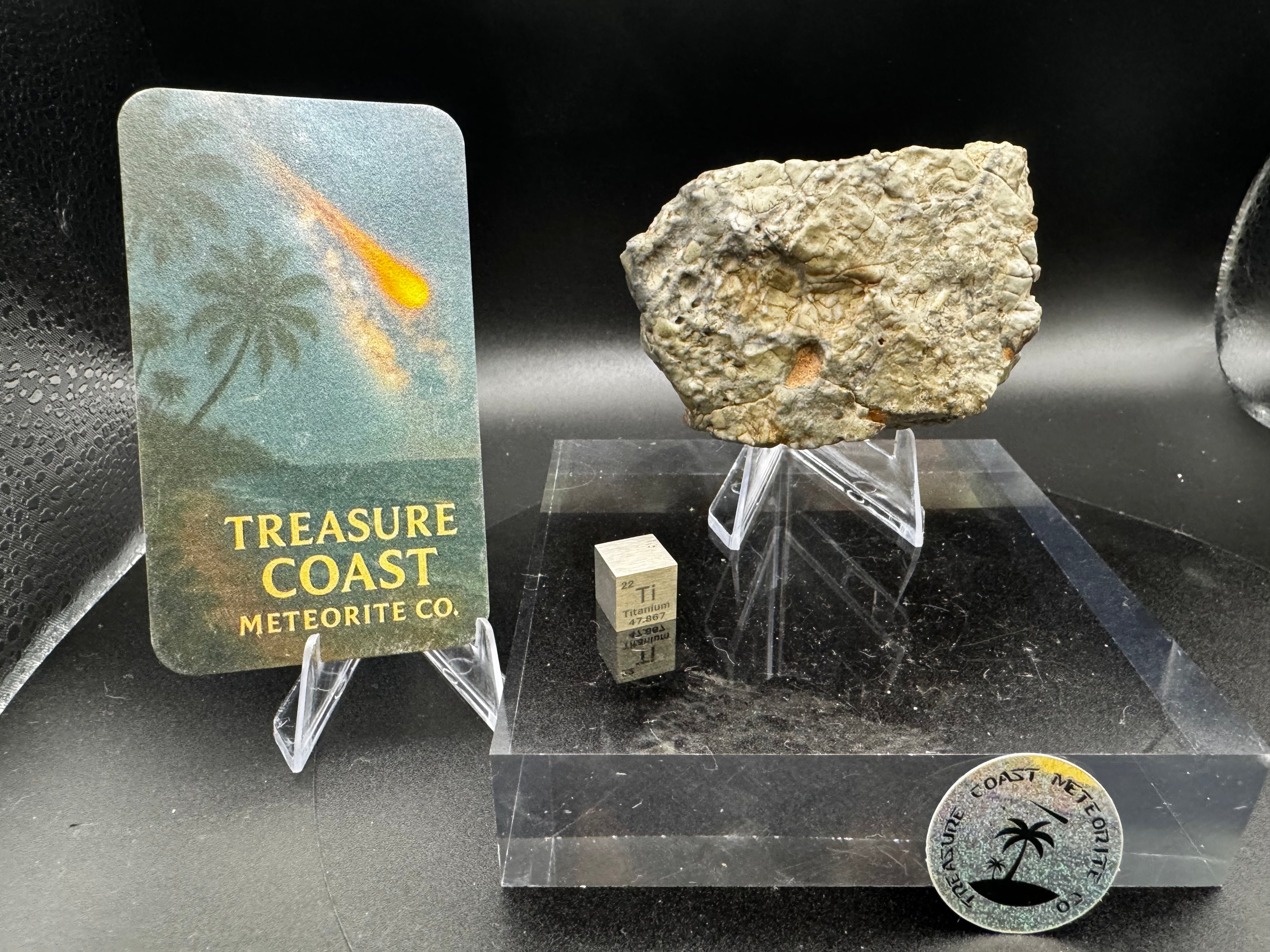 Adrar 013 lunar meteorite 142.20g on display stand with Treasure Coast Meteorite Co. branded card and meteorite cube for scale