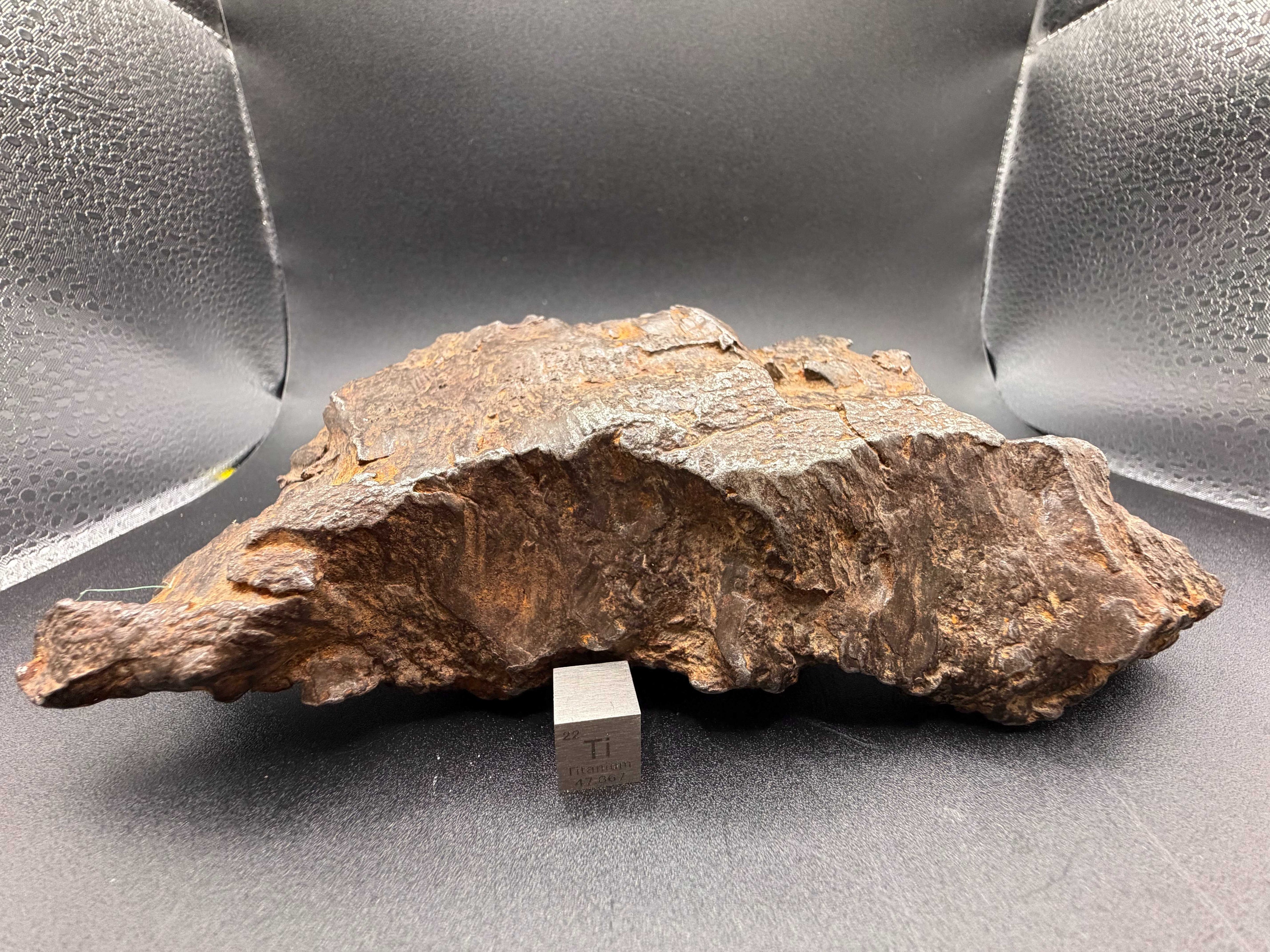 Gebel Kamil 1414g iron meteorite displayed with a titanium scale cube, showing the broad sheared face
