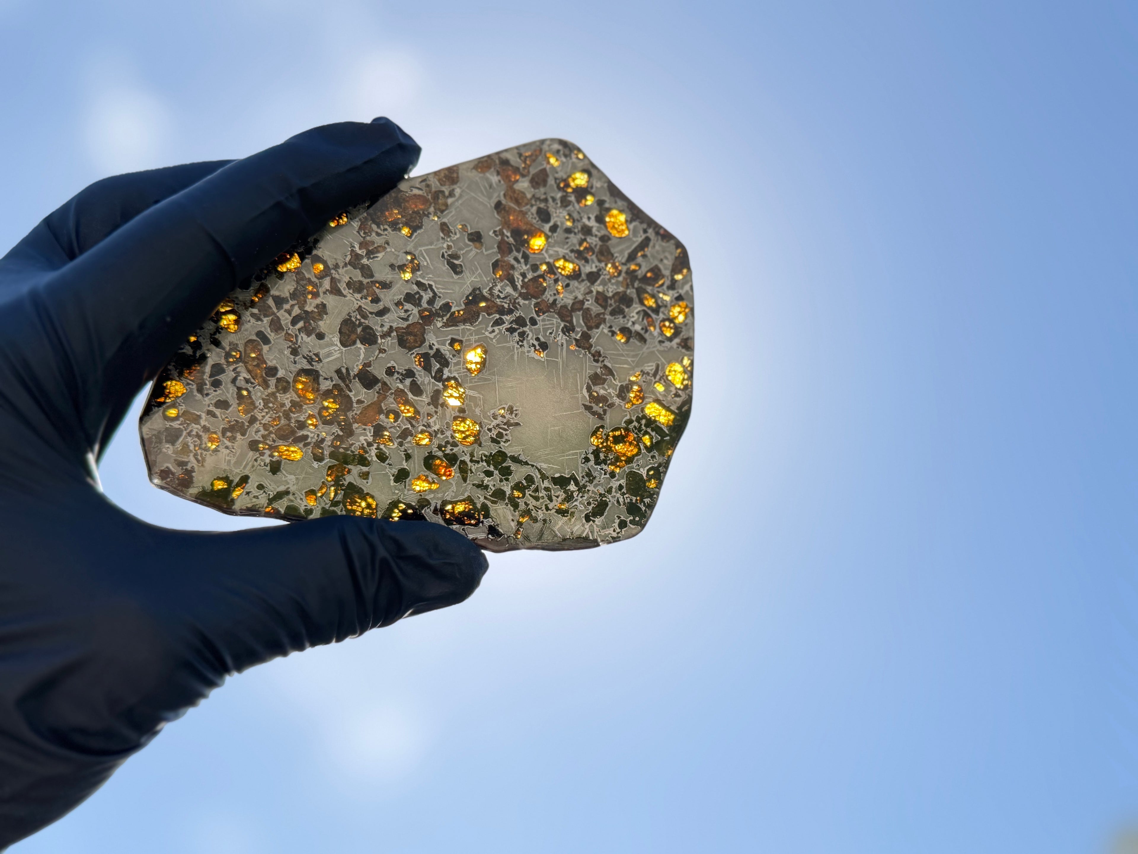 Backlit hand-held view, Gyarub Zangbo Pallasite, translucent olivine crystals lit by daylight against blue sky, 86.11g