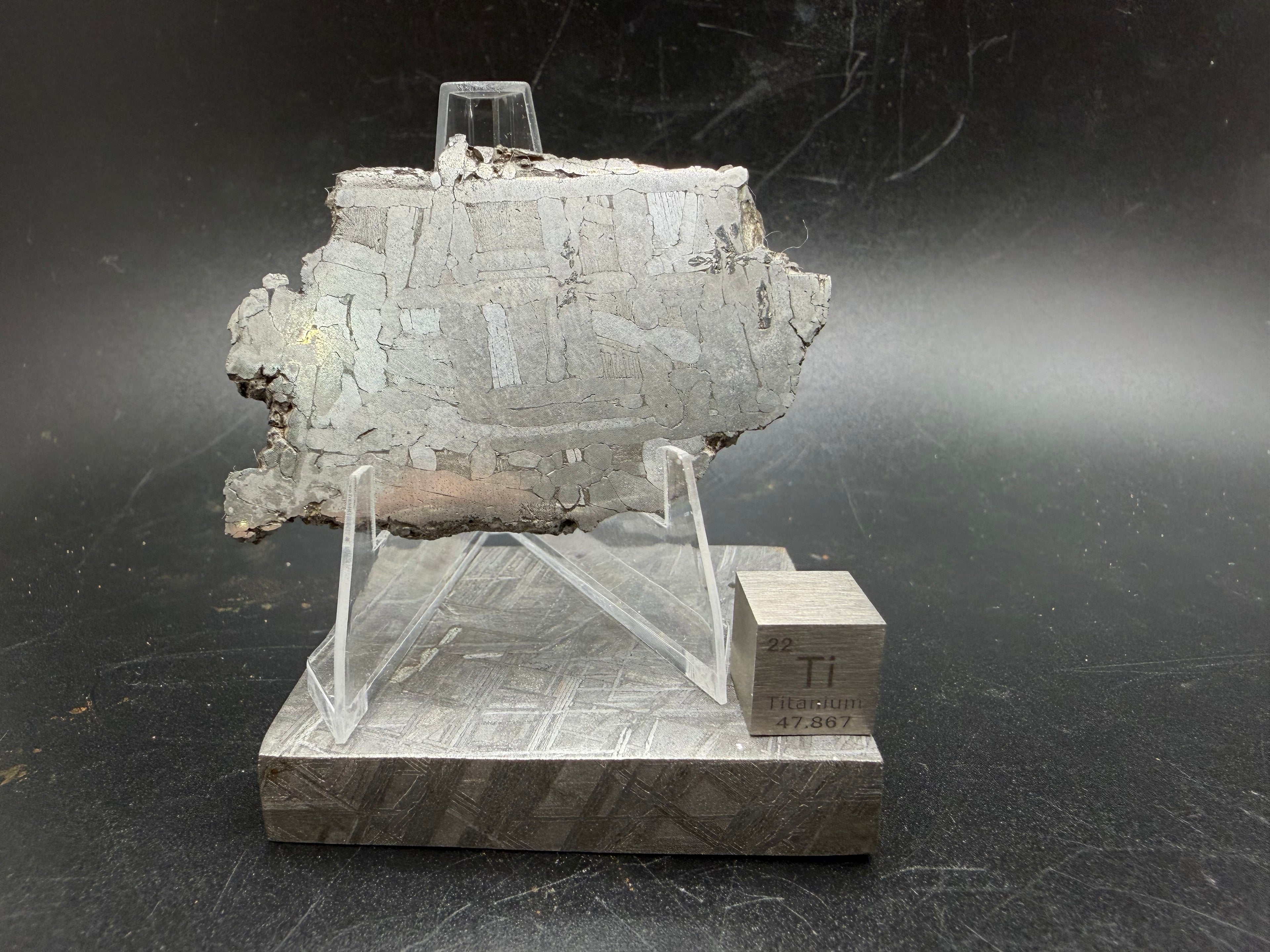 Kaalijärv iron meteorite 29.51g full slice on acrylic display stand front view showing Widmanstätten pattern with heavy weathered crust on edges and titanium cube for scale