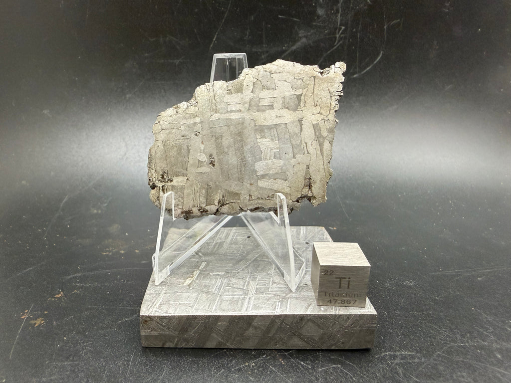 Kaalijärv iron meteorite 33.00g full slice on acrylic display stand front view showing Widmanstätten pattern weathered natural edge and titanium cube for scale