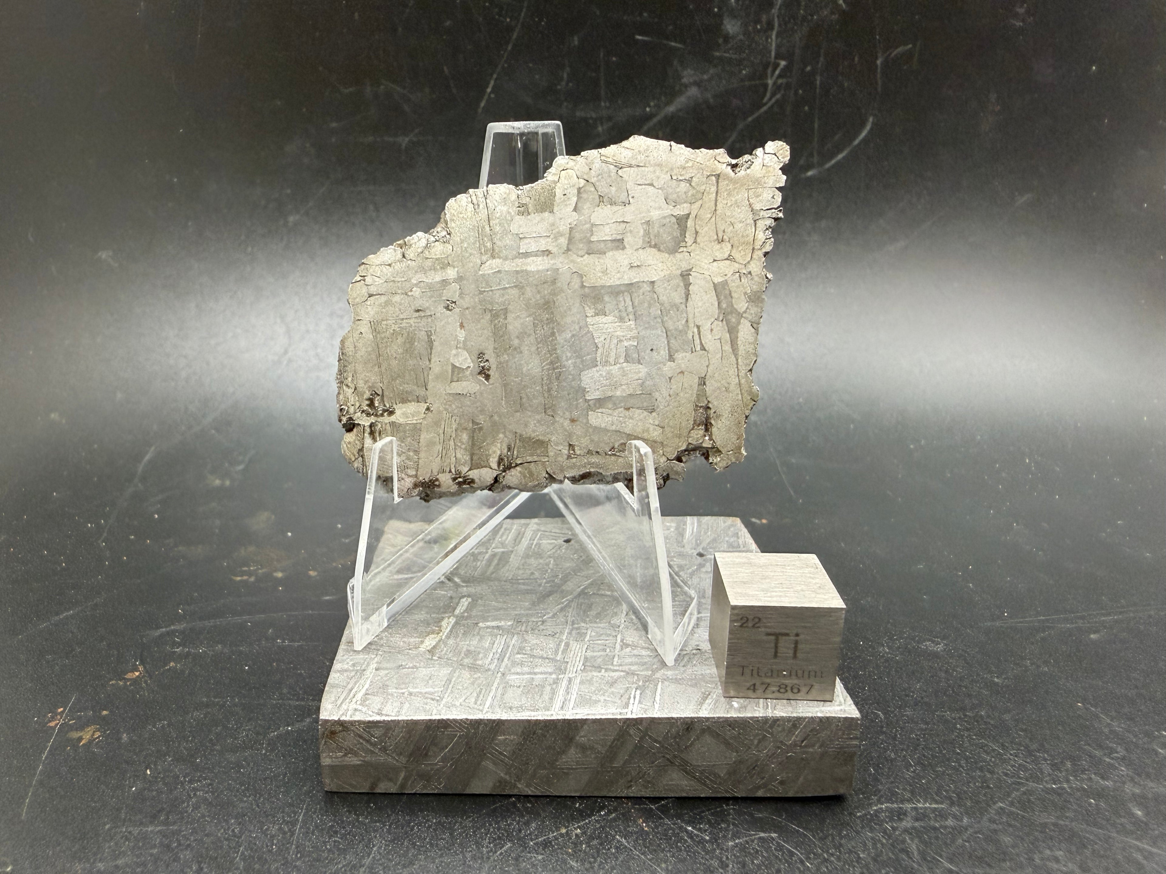 Kaalijärv iron meteorite 33.00g full slice on acrylic display stand front view showing Widmanstätten pattern weathered natural edge and titanium cube for scale
