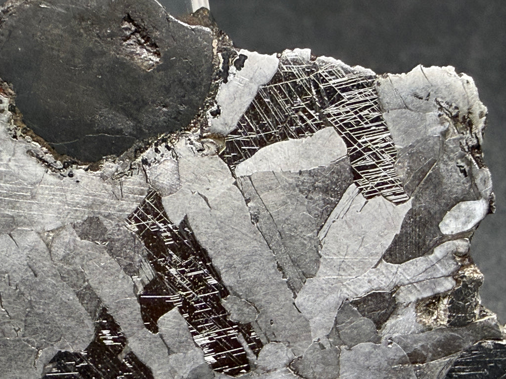 Kaalijarv iron meteorite, IAB-MG, 15.78 g slice, close-up of the rounded dark inclusion and the surrounding etched metal, with Neumann bands crossing the darker zones