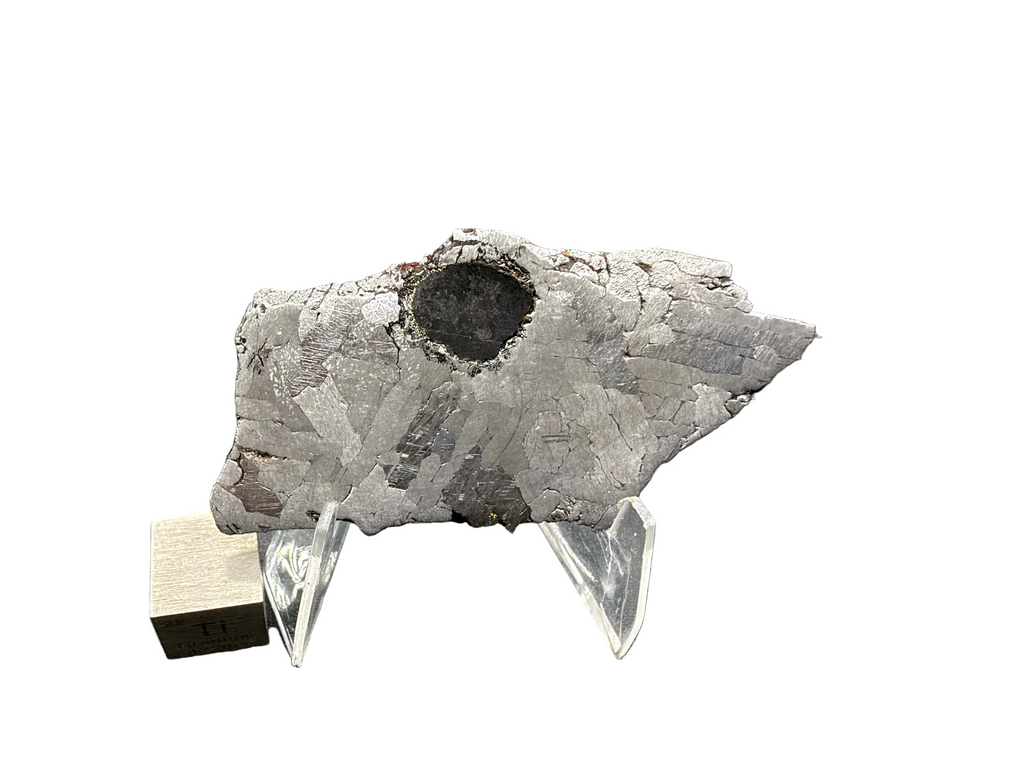 Kaalijarv IAB-MG iron meteorite slice, 17.00 g, front face etched, on an acrylic stand with a large rounded dark inclusion near the top edge and a titanium cube for scale