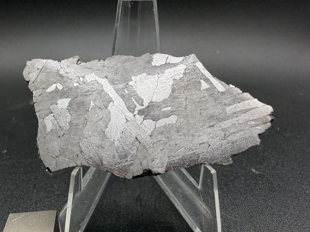 Kaalijarv iron meteorite, IAB-MG, 21.00 g slice on a clear acrylic stand with a scale cube, etched face lit from another angle so the Widmanstatten plates appear bright against gray
