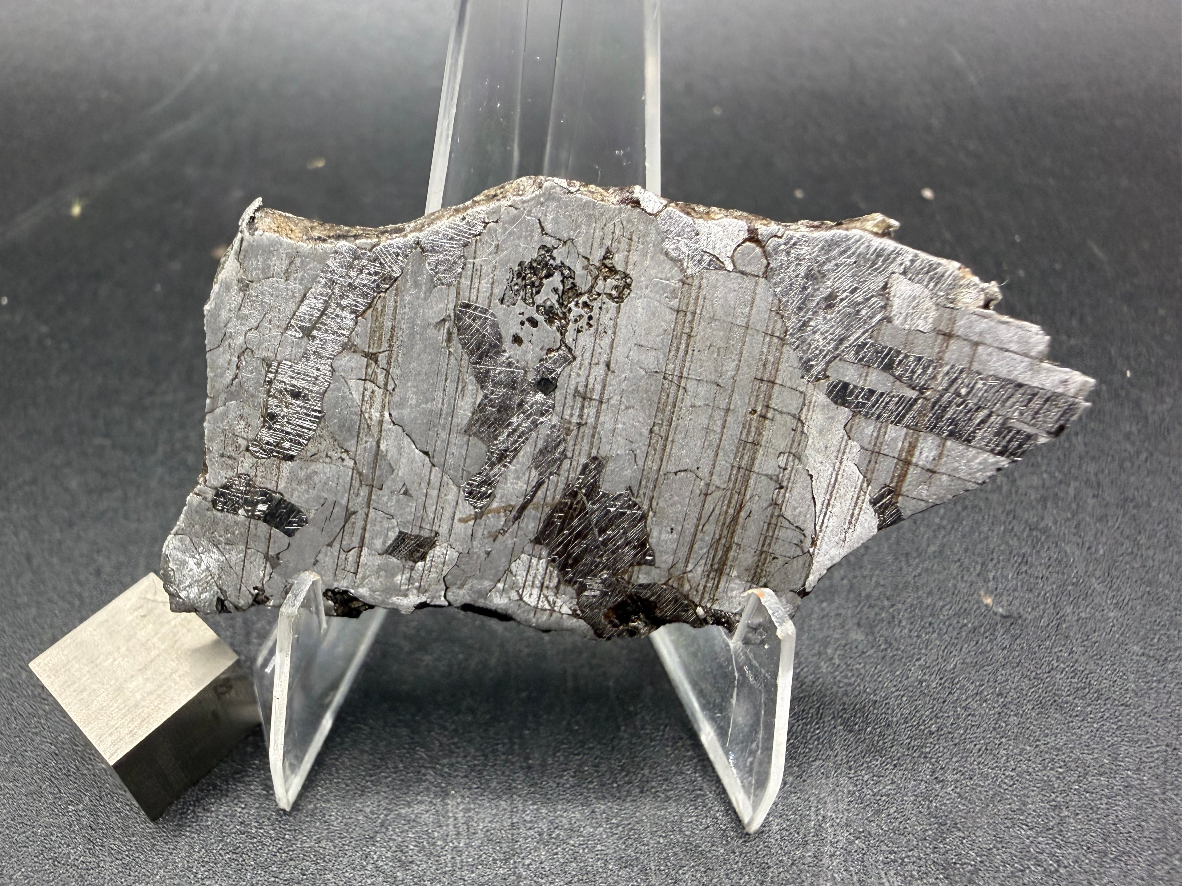 Kaalijarv iron meteorite, IAB-MG, 21.00 g slice on a clear acrylic stand with a scale cube, etched face with fine brown streaks, darker plates showing Neumann bands, and a small cluster of dark pits near the top edge
