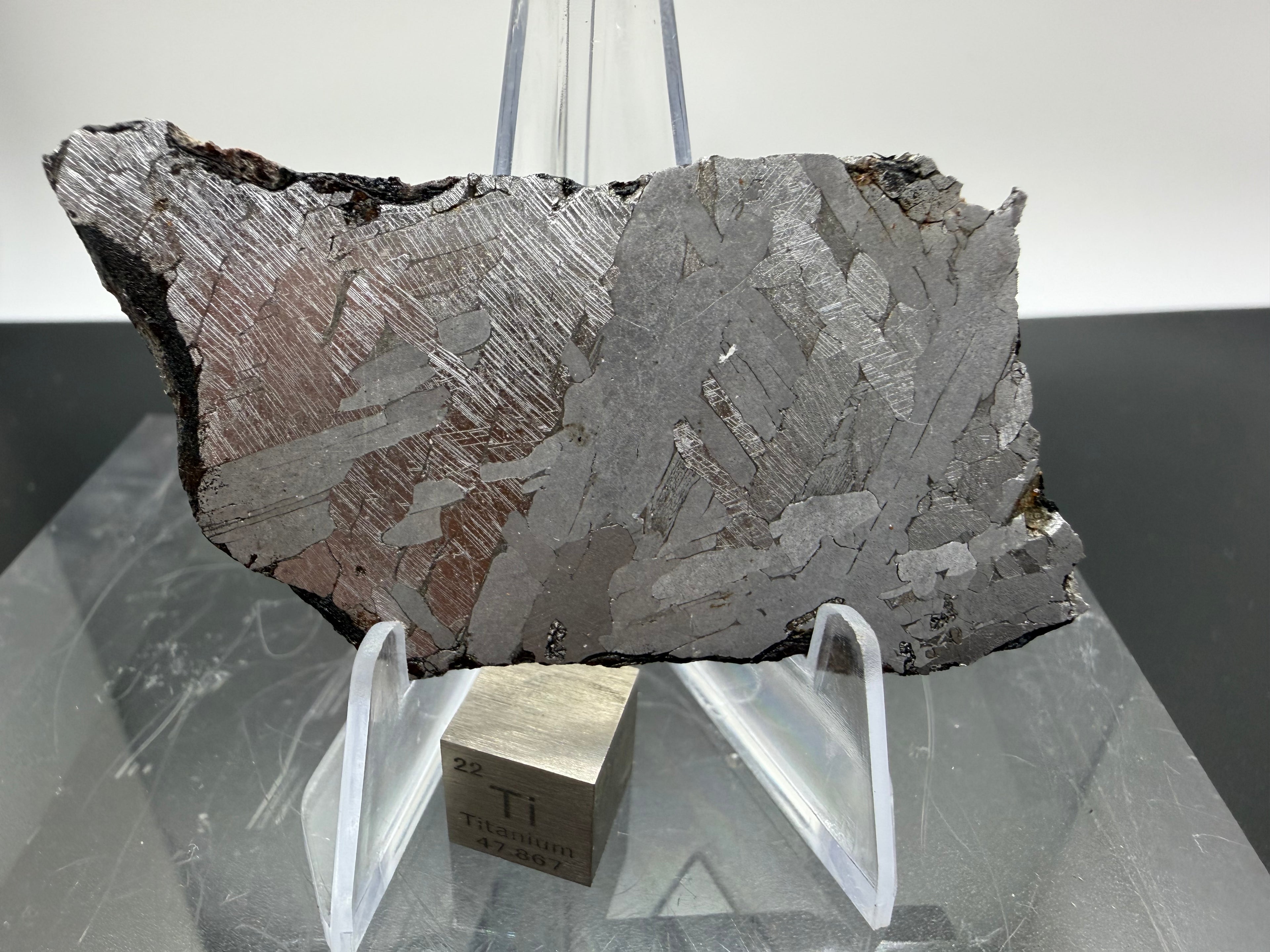 Kaalijarv iron meteorite, IAB-MG, 27.00 g slice on a clear acrylic stand with a scale cube, full view of the etched face showing crossing plates with Neumann bands and a weathered edge