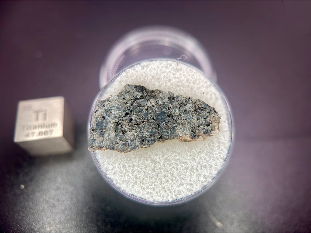 Capsule display of Ksar Ghilane 022 ungrouped achondrite meteorite slice showing dark pyroxene-rich matrix, 1.75g
