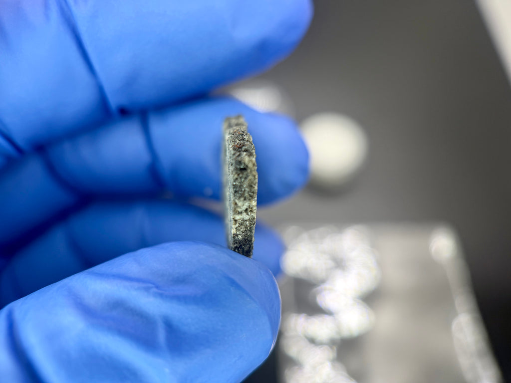 Edge profile of Ksar Ghilane 022 ungrouped achondrite meteorite slice held in gloved hand showing thickness and pyroxene-dominated cross-section, 1.75g