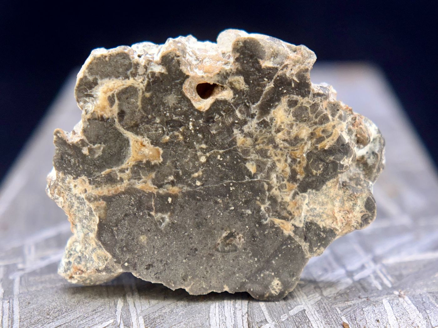NWA 17706 lunar meteorite slice, 2.00g, troctolitic anorthosite melt breccia showing gray matrix with tan veining and a small vug, displayed on an etched iron stand