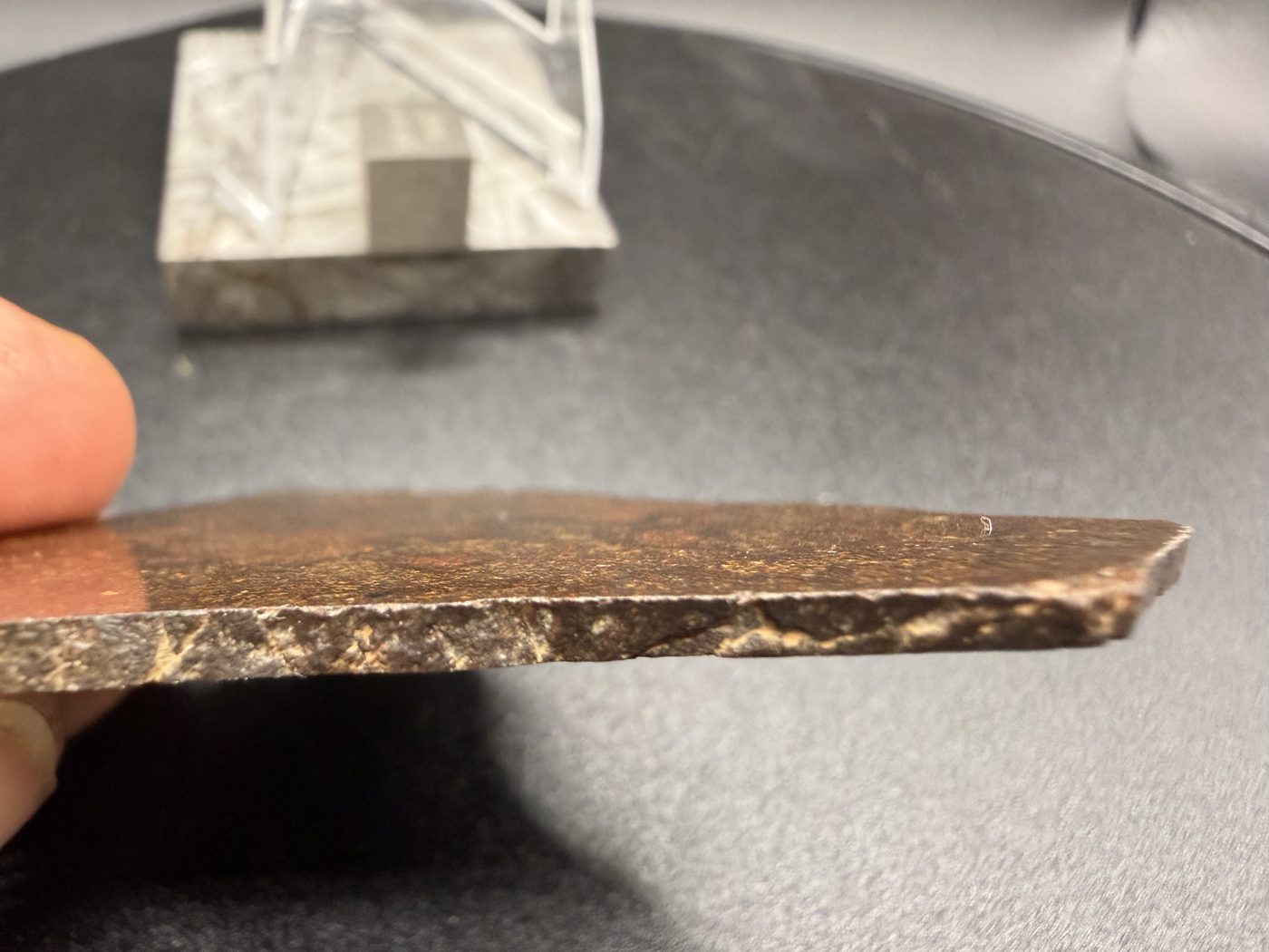 Edge profile of NWA 17916 CO3 carbonaceous chondrite meteorite slice held in hand showing thickness and pale caliche veining along the edge, 12.00g